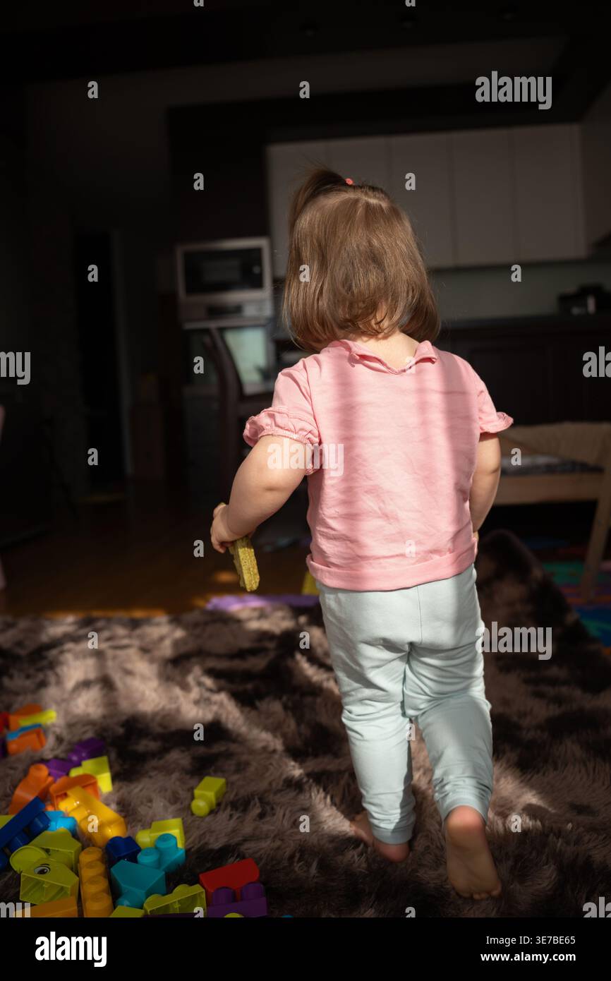 vista posteriore di un bambino con una camicia rosa che regge un blocco di giocattoli, cammina a piedi nudi su un morbido tappeto marrone in un interno illuminato dal sole del soggiorno e della cucina Foto Stock