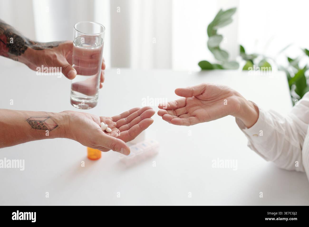 Mani di un figlio di razza mista che dà pillole e un bicchiere d'acqua alla sua madre anziana mentre è seduto a tavola nel salotto di casa Foto Stock