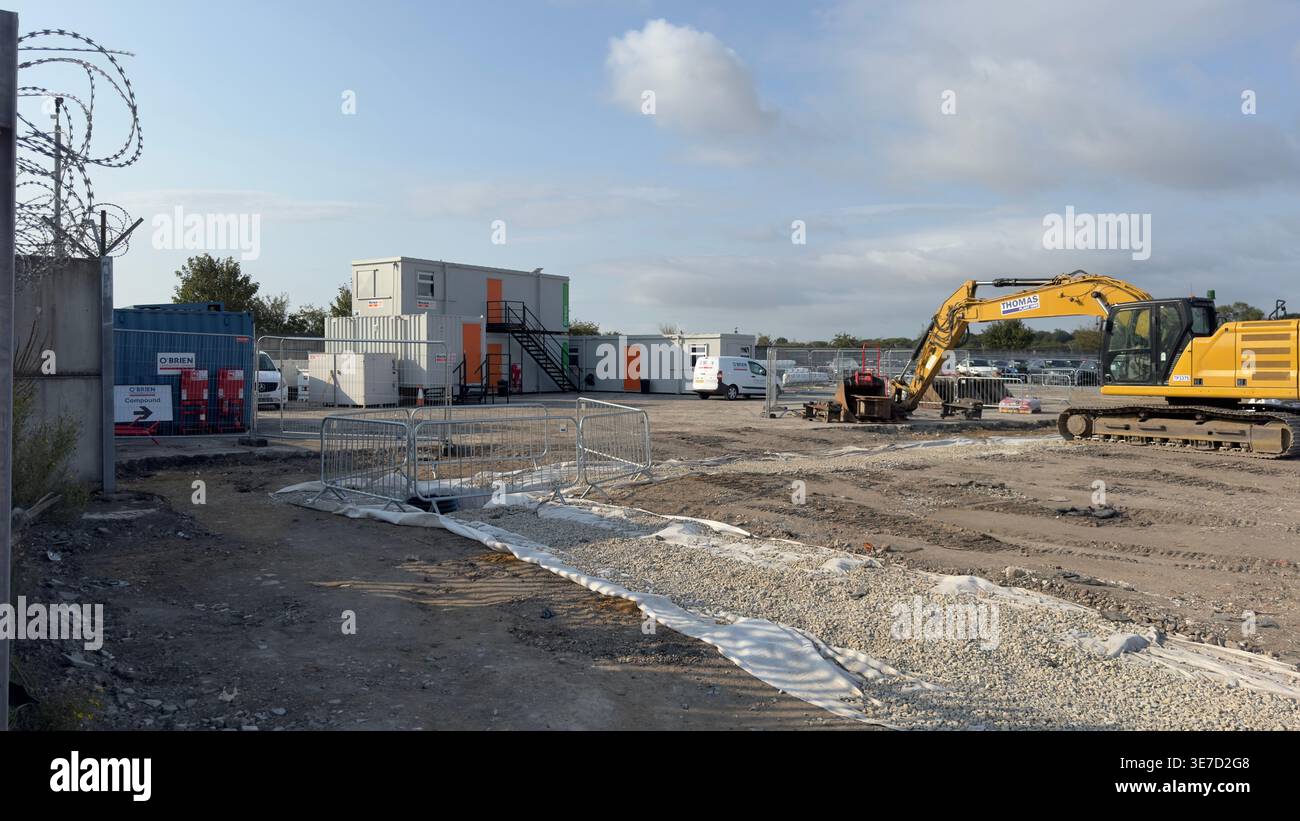 Escavatore giallo parcheggiato vicino a edifici modulari temporanei in un cantiere attivo con recinzione e ghiaia a Copart, York. Foto Stock