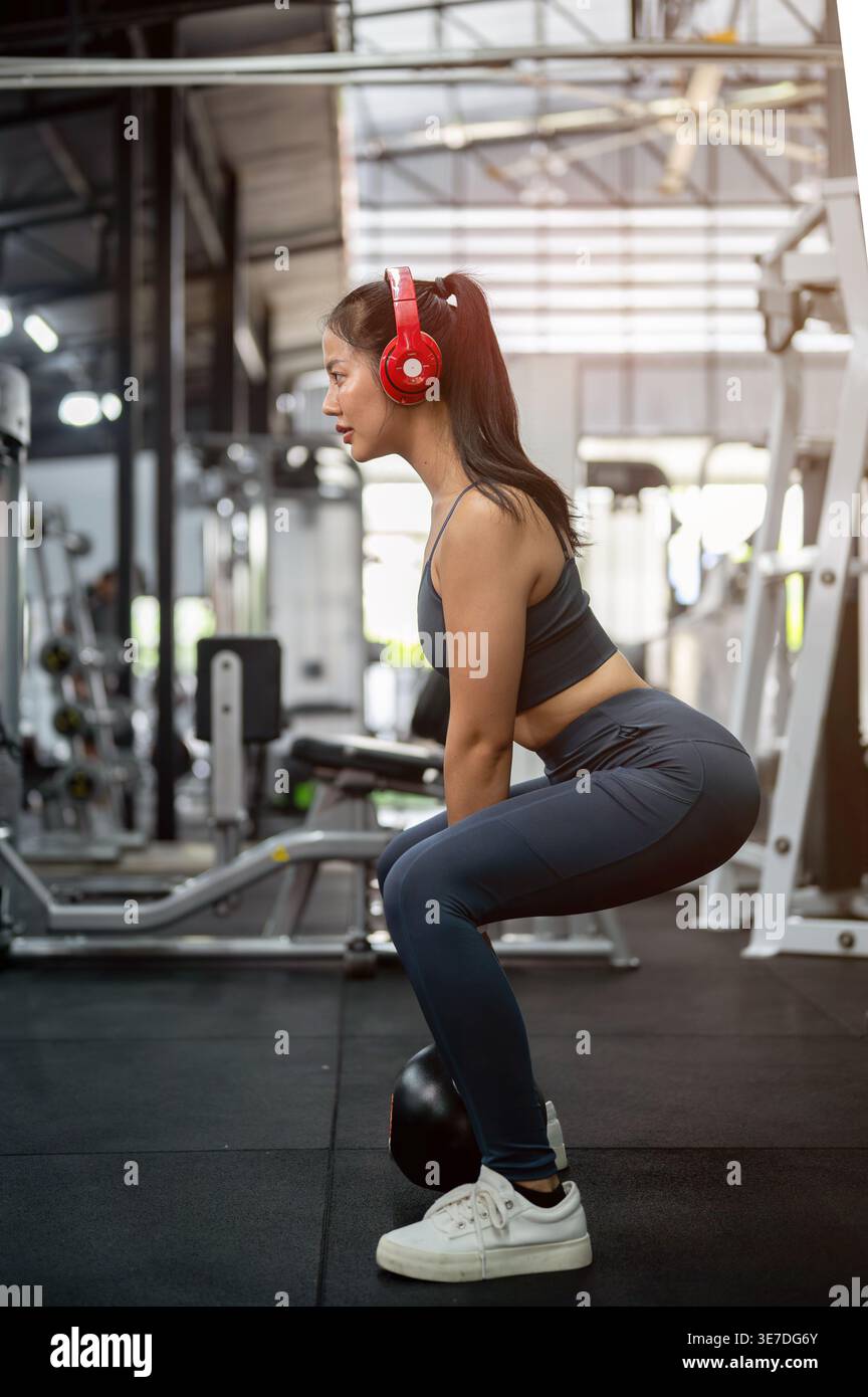 La donna thailandese in forma fisica si veste ascoltando la musica con le cuffie e guardando lontano mentre esegue gli squat kettlebell in palestra Foto Stock