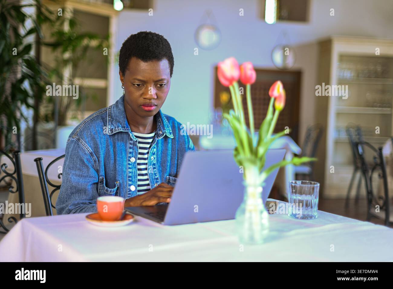 Donna nera con i capelli corti che indossa una giacca in denim che lavora su un computer portatile mentre è seduta al tavolo del ristorante Foto Stock