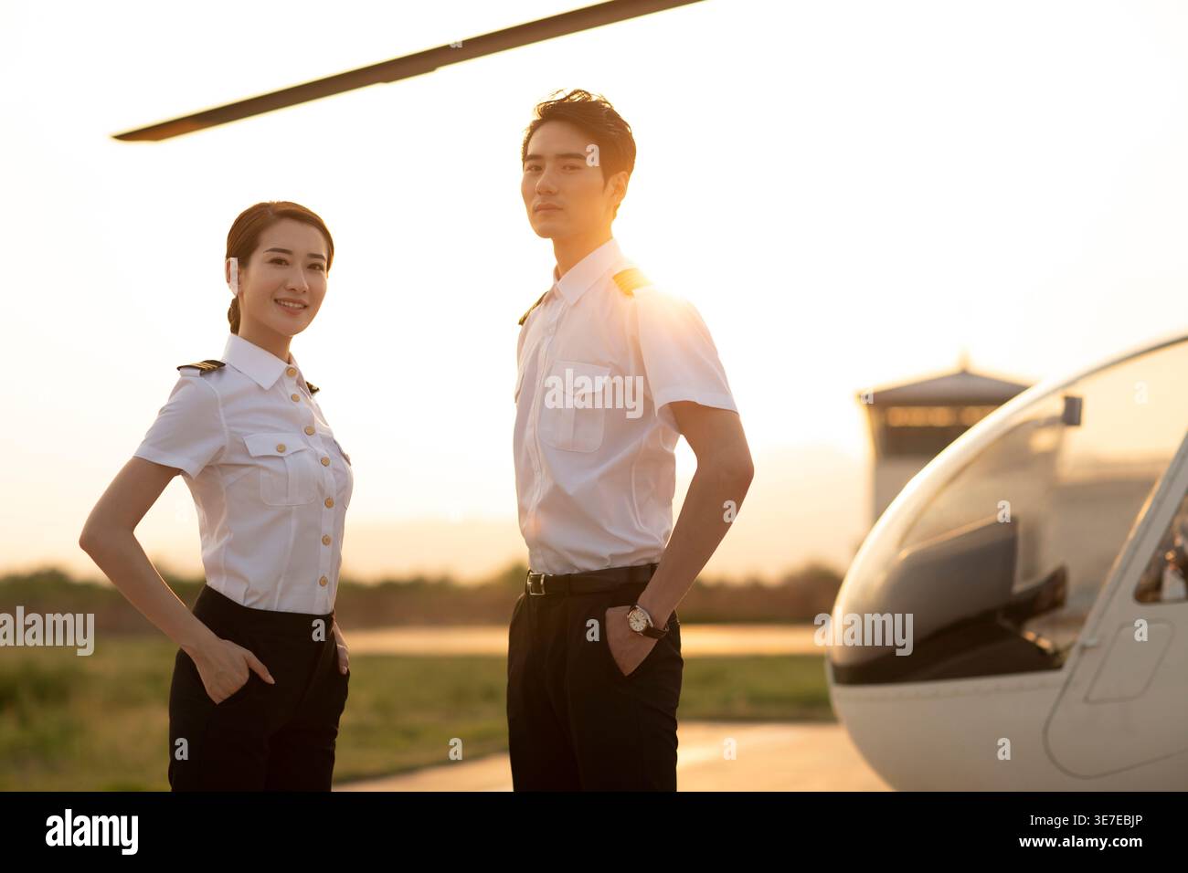 Piloti cinesi sorridenti uomini e donne in piedi con le mani in tasca vicino all'elicottero durante il tramonto Foto Stock