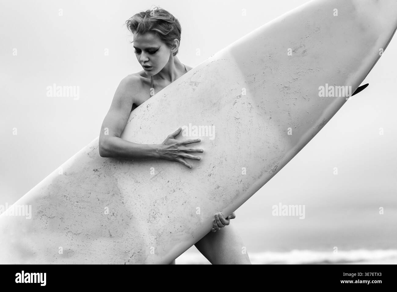 Ritratto monocromatico della donna FIT con capelli eleganti e trucco che regge la tavola da surf. La composizione diagonale e la luce soffusa creano un'atmosfera suggestiva e penosa. Co Foto Stock