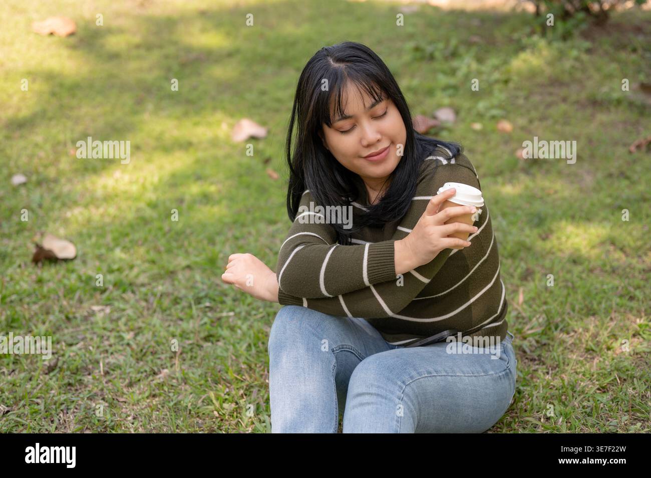 Donna asiatica che tiene una tazza di caffè braccia incrociate gomito appoggiato sul ginocchio mentre siede sull'erba con gli occhi chiusi nel parco Foto Stock
