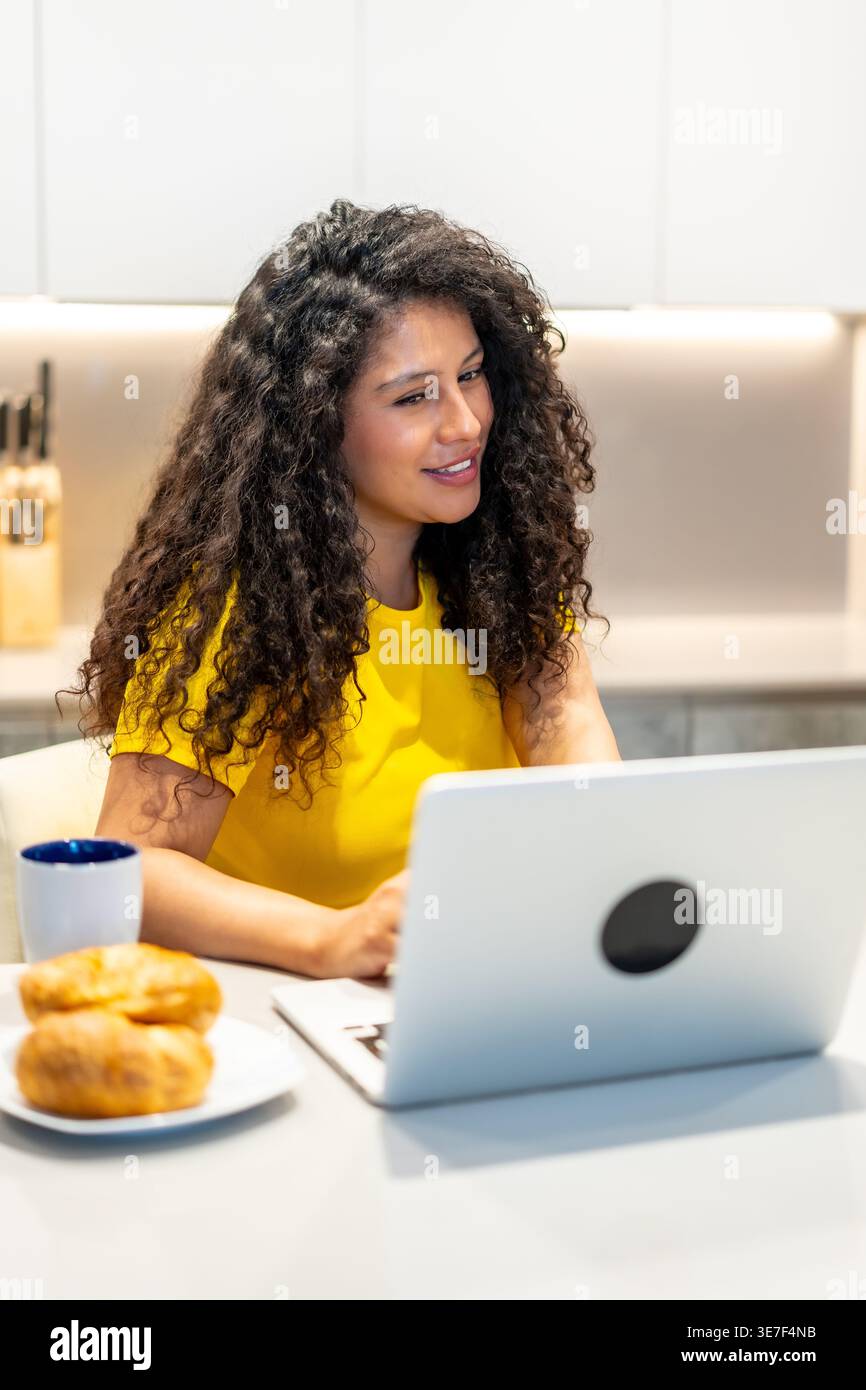 Donna ispanica con capelli ricci che partecipa a una riunione online utilizzando un computer portatile mentre fa colazione al bancone della cucina a casa Foto Stock