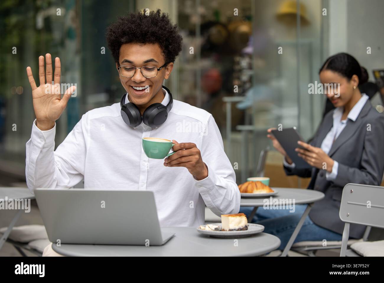 Uomo nero che indossa occhiali da vista e cuffie sul collo che ondeggia la mano bevendo un caffè e facendo videochiamata sul laptop mentre si siede al tavolo vicino a una donna... Foto Stock