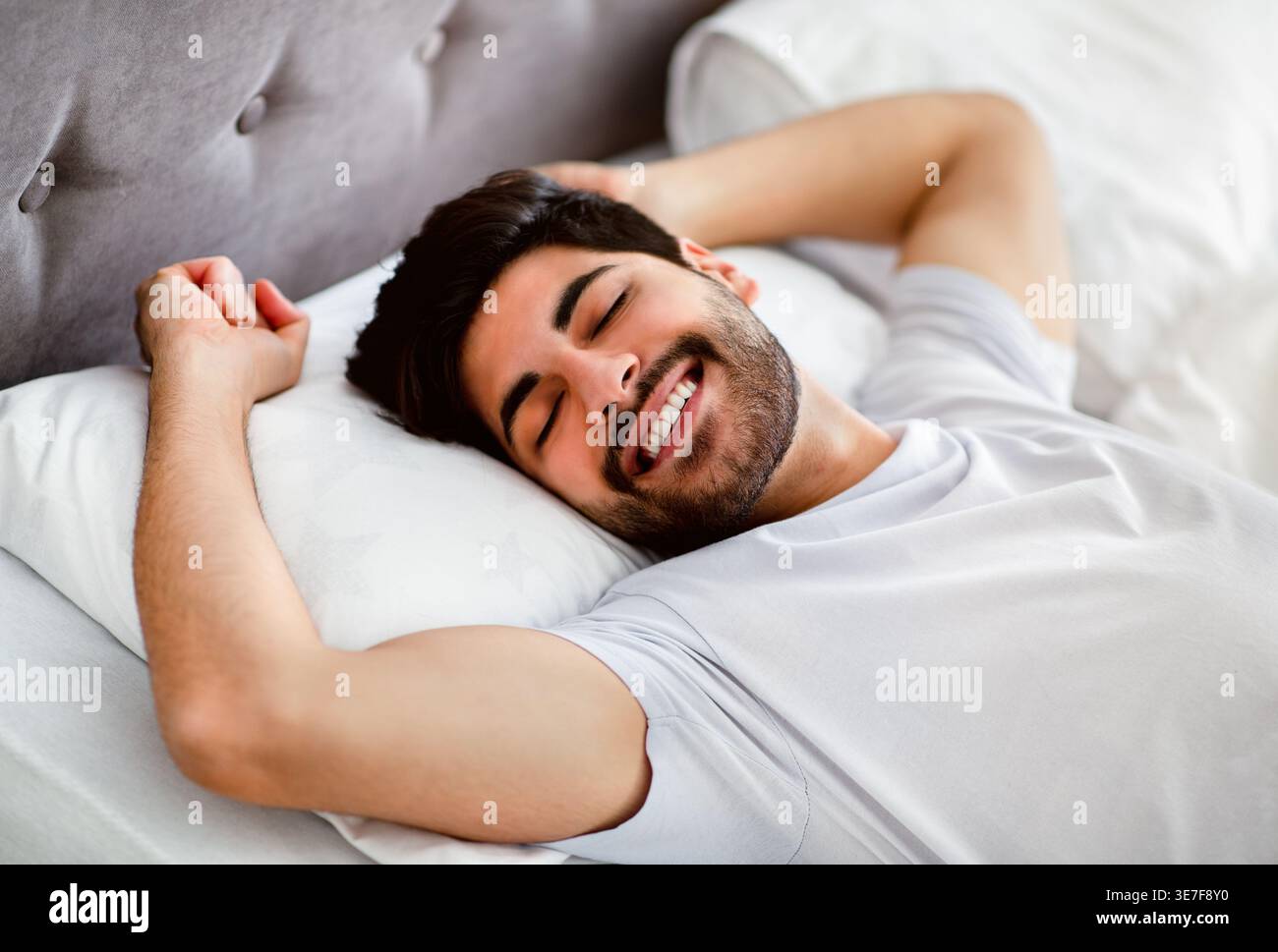 Uomo arabo sorridente con la barba che indossa una camicia bianca che allunga le braccia con gli occhi chiusi mentre è sdraiato sul letto in camera da letto di casa Foto Stock