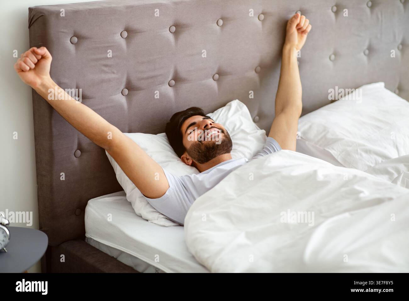 Uomo arabo sorridente con la barba che indossa una camicia bianca che allunga le braccia con gli occhi chiusi mentre è sdraiato sul letto in camera da letto di casa Foto Stock