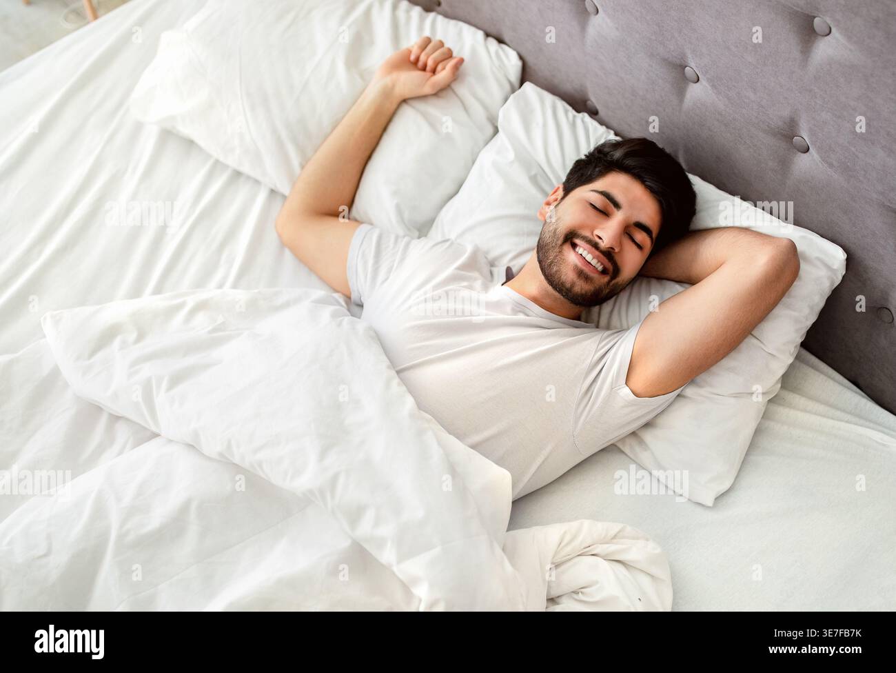 Uomo arabo sorridente con la barba che indossa una camicia bianca che allunga le braccia con gli occhi chiusi mentre è sdraiato sul letto in camera da letto di casa Foto Stock
