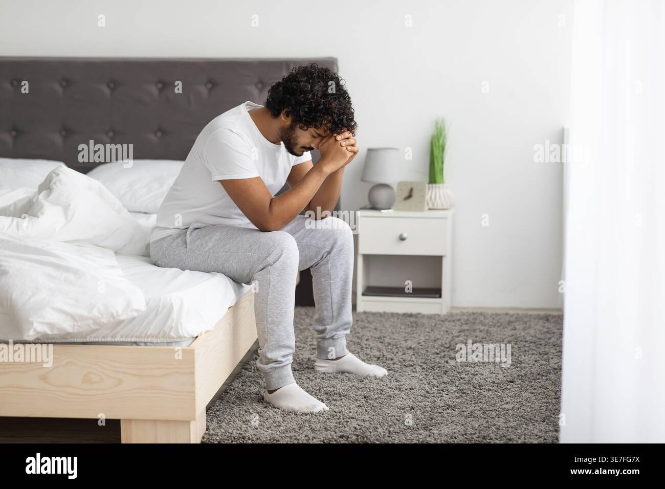 Uomo indiano barbuto stressato con capelli ricci appoggiati sulla fronte su pugni clenati seduti sul letto in camera da letto di casa Foto Stock