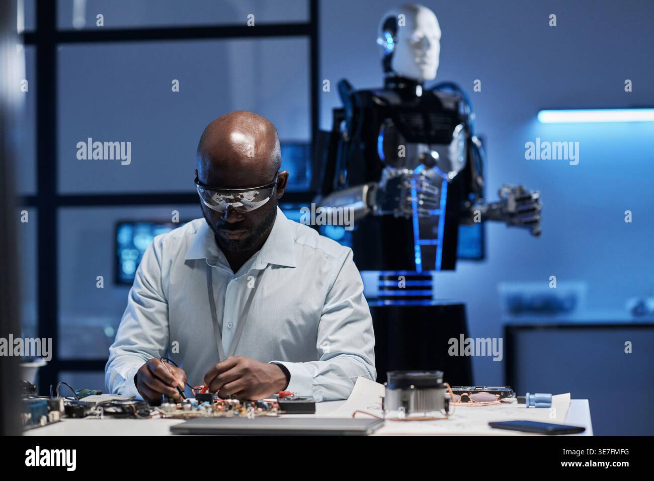 Ingegnere informatico maschio nero in camice da laboratorio e occhiali protettivi che lavorano sulla scheda madre seduto al tavolo di fronte al robot in laboratorio Foto Stock