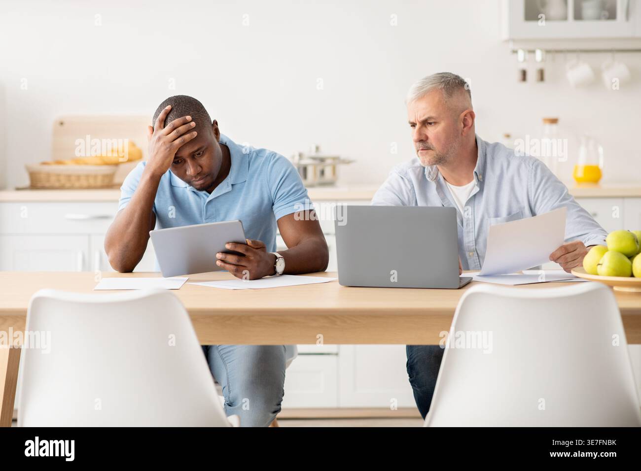 Uomo nero stressato che si appoggia alla testa e usa il tablet seduto a tavola con un amico di mezza età caucasico che lavora su documenti e grembo... Foto Stock