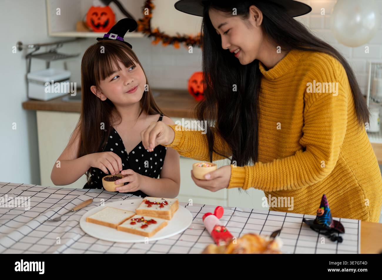 Madre e figlia asiatica sorridenti che indossano cappelli da strega che preparano il cibo per Halloween mentre sono seduti a tavola in cucina a casa Foto Stock