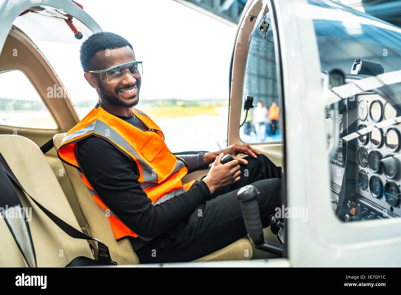 Un ingegnere afroamericano maschio sorridente che siede all'interno di un jet privato che tiene il controllo sulla cabina di pilotaggio nell'hangar Foto Stock