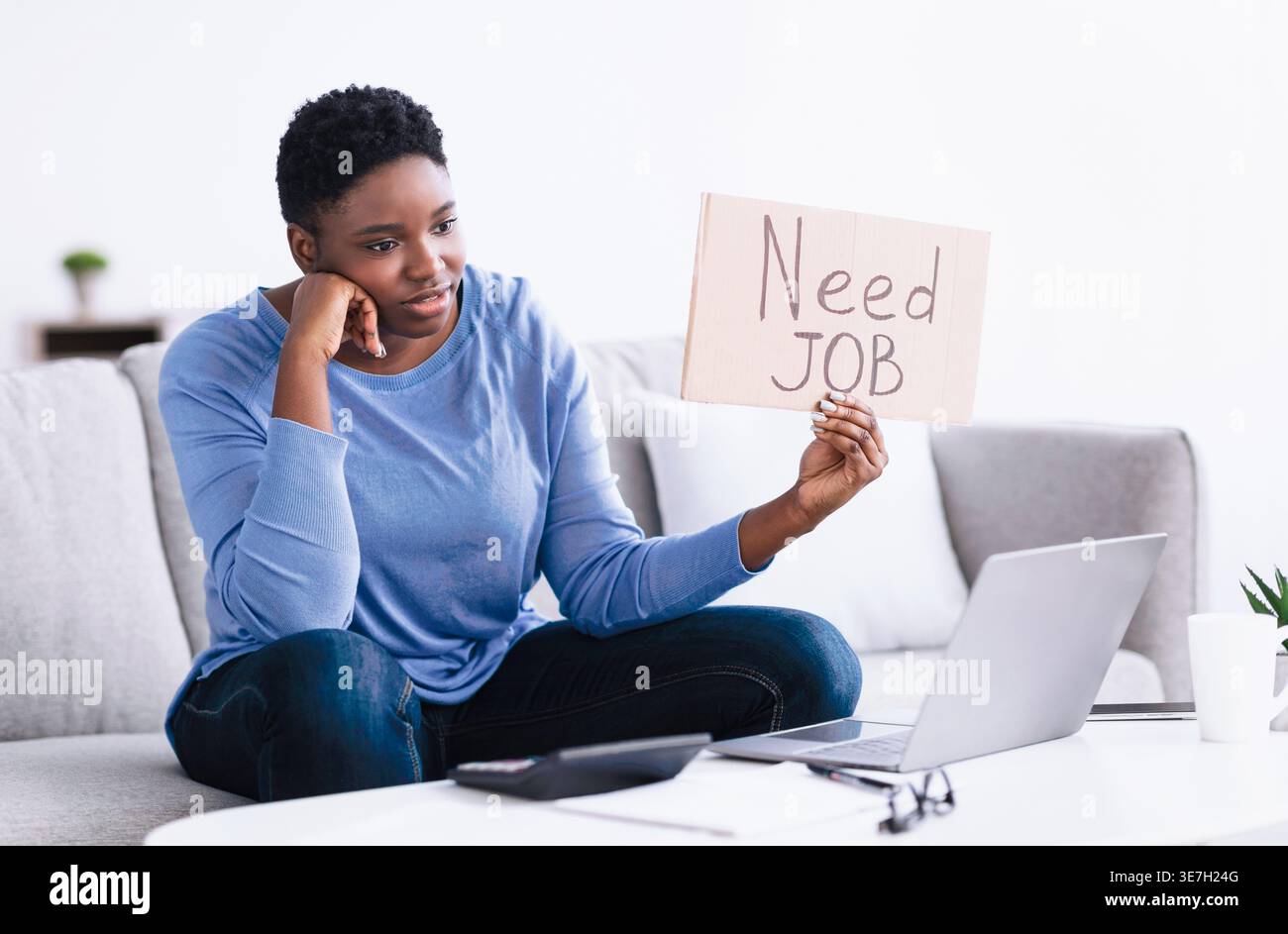 Donna nera con afro corto che tiene in mano un cartello di cartone con il bisogno di un lavoro scritto testo mentre si sta videochiamando attraverso il computer portatile in salotto di casa Foto Stock