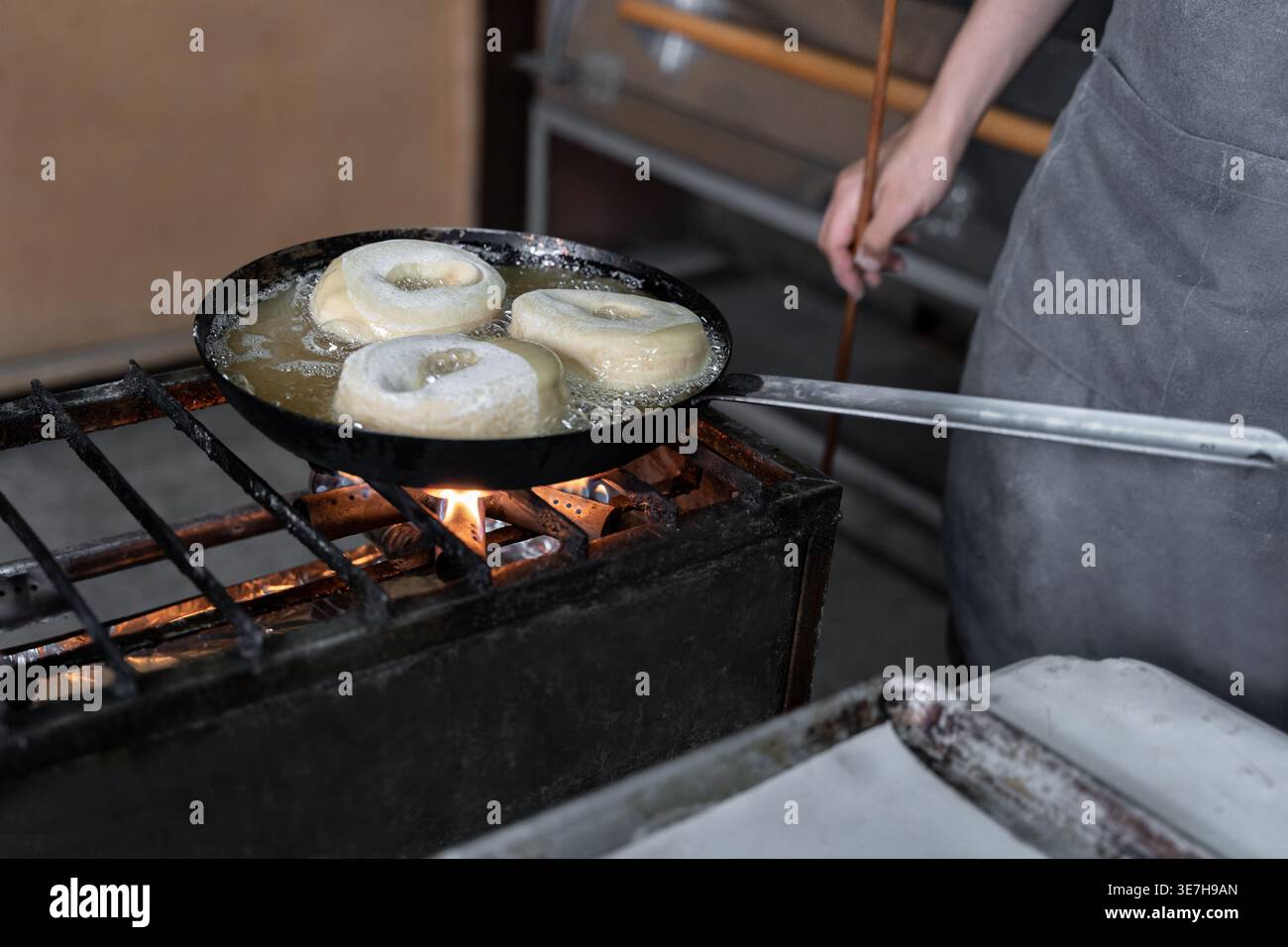 panettiera ispanica in grembiule in piedi a tavola e friggere ciambelle in fornelli a gas con bastoncino in panetteria Foto Stock