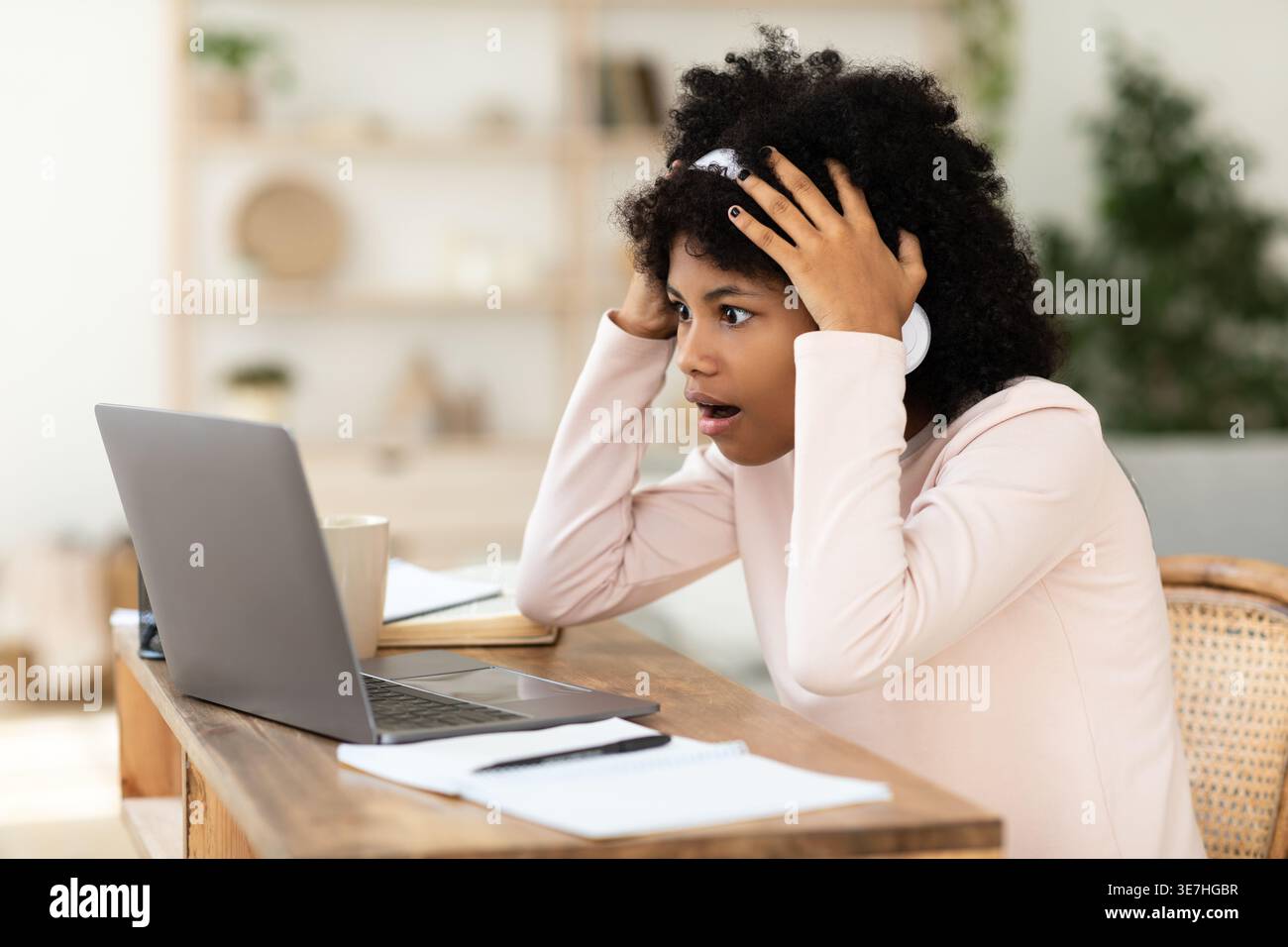 Ragazza preadolescente nera scioccata con afro che indossa le cuffie che guarda il laptop seduto alla scrivania in salotto Foto Stock