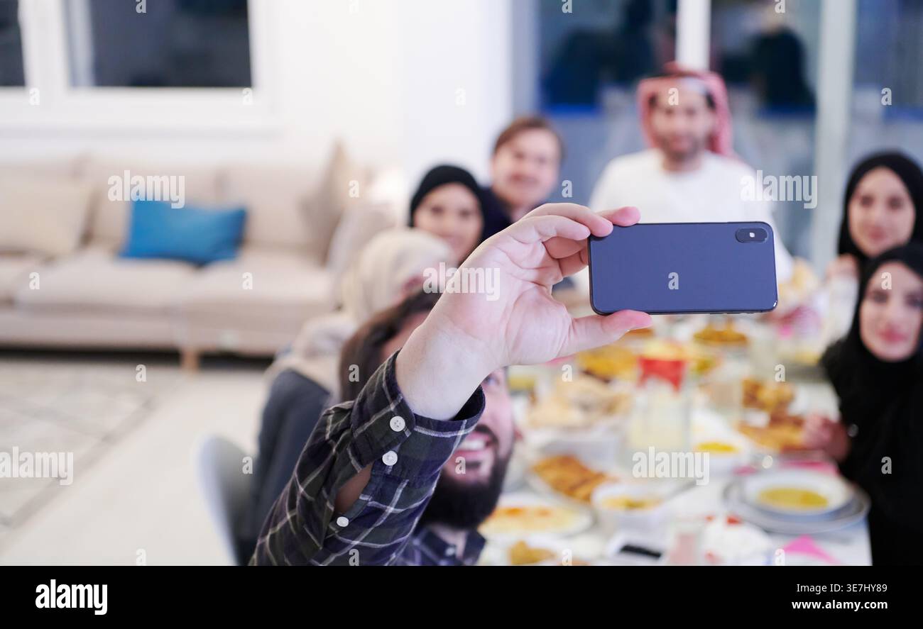 Famiglia musulmana sorridente seduta a tavola a scattare selfie sullo smartphone prima di cenare Iftar durante il mese di festa del Ramadan a casa Foto Stock