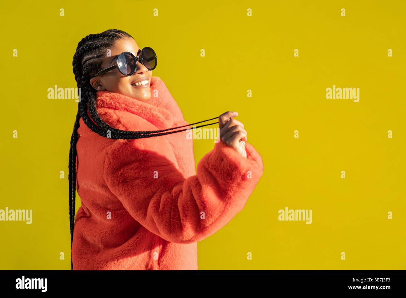 Giovane donna afroamericana con occhiali da sole trecce e cappotto arancione, con sfondo colorato Foto Stock