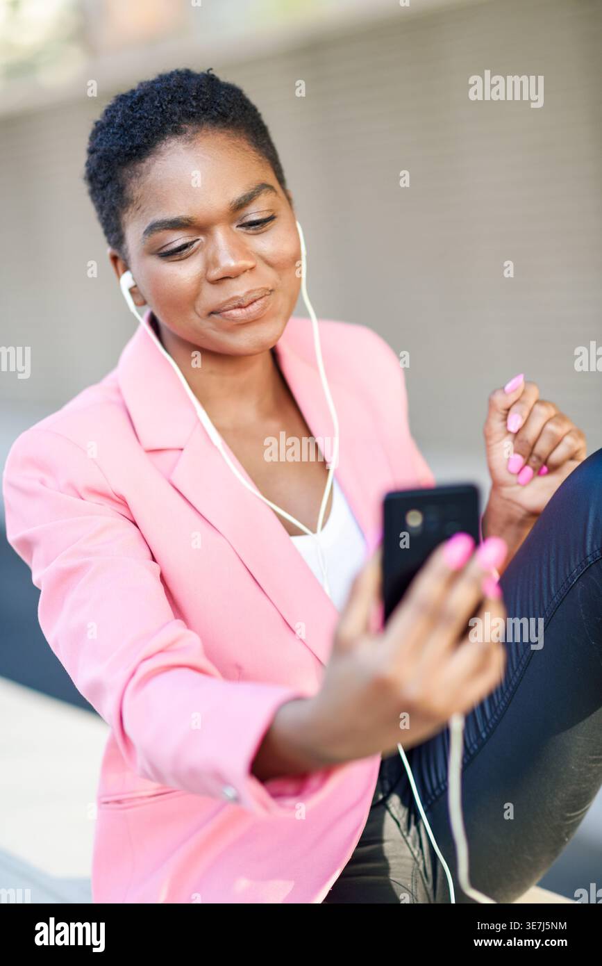 Donna d'affari nera sorridente in costume rosa che effettua videochiamate con il suo smartphone mentre è seduta all'aperto Foto Stock