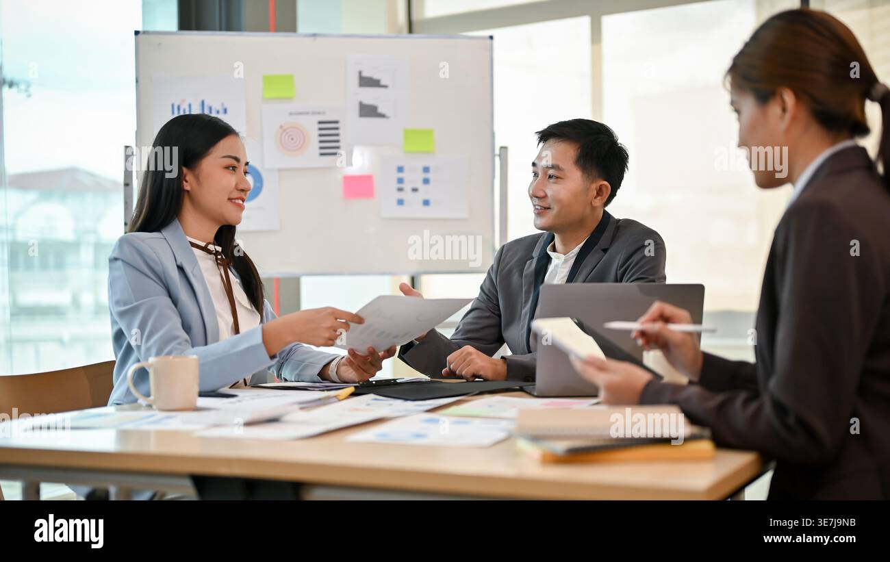 Uomo d'affari asiatico sorridente che lavora insieme a colleghi seduti a tavola e che discute relazioni finanziarie in ufficio Foto Stock