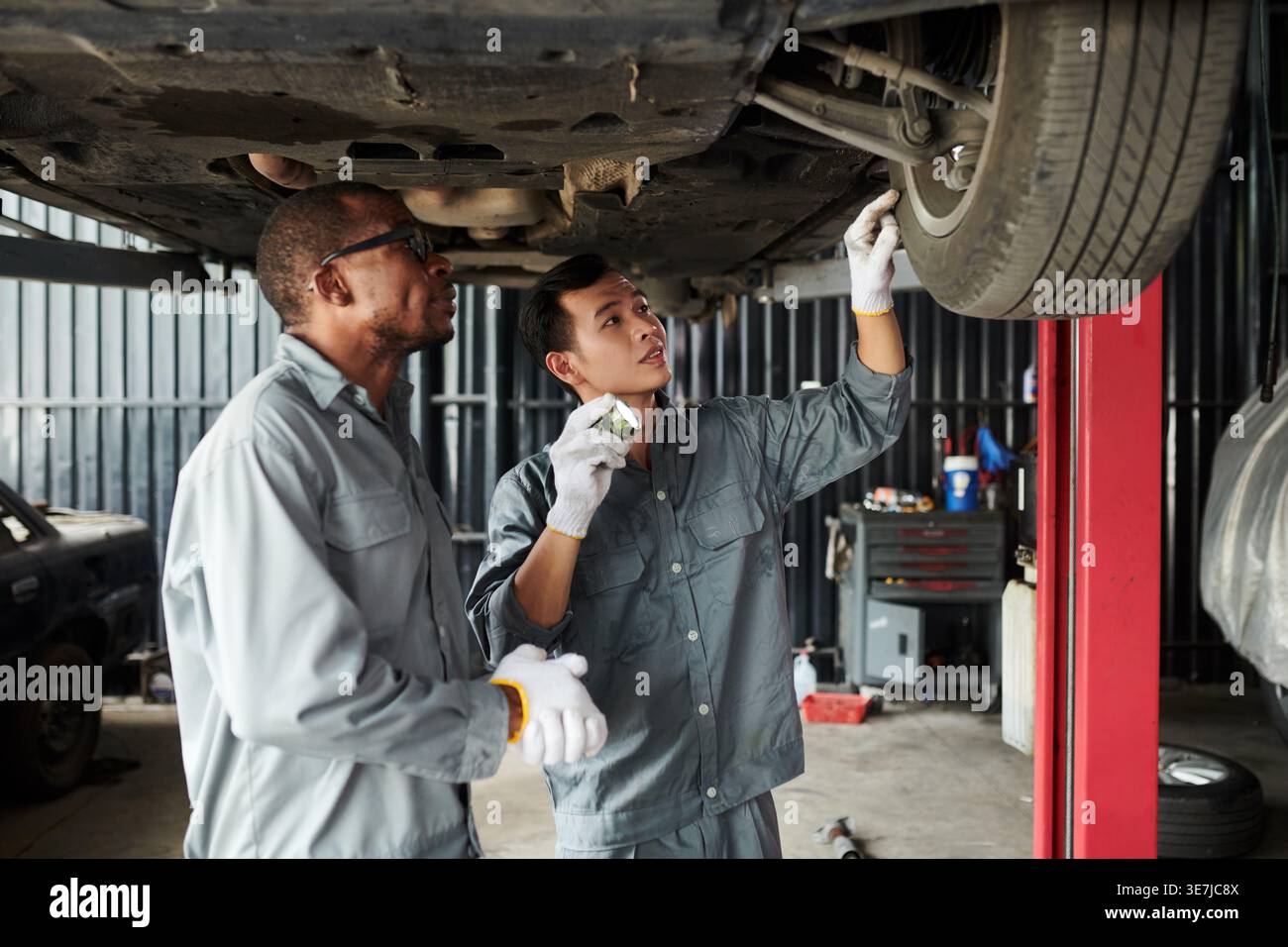 Meccanici maschi neri e asiatici che indossano guanti che riparano sotto il telaio dell'auto mentre si trova in un'officina di riparazione auto Foto Stock