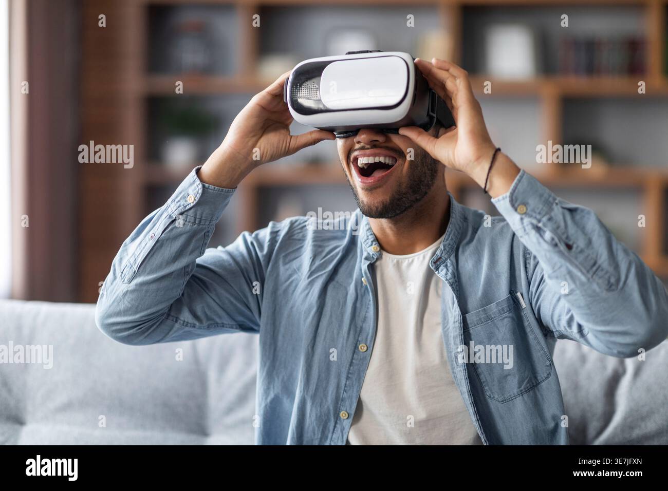 Uomo nero che indossa occhiali VR e goditi un'esperienza di realtà virtuale seduto sul divano di casa Foto Stock