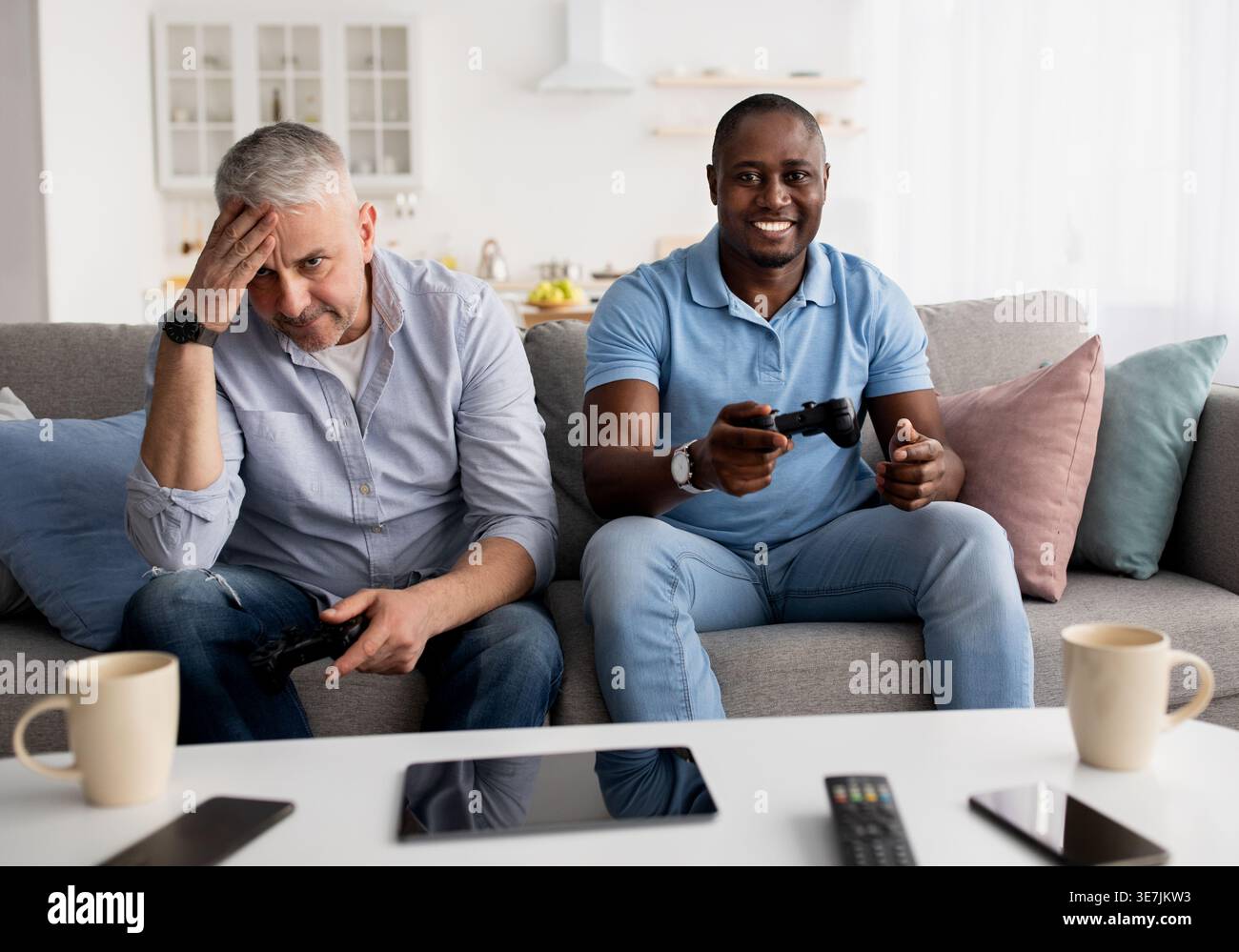 Uomo nero e amico di mezza età caucasico maschio che si diverte a giocare con i controller mentre si siede sul divano nel soggiorno di casa Foto Stock