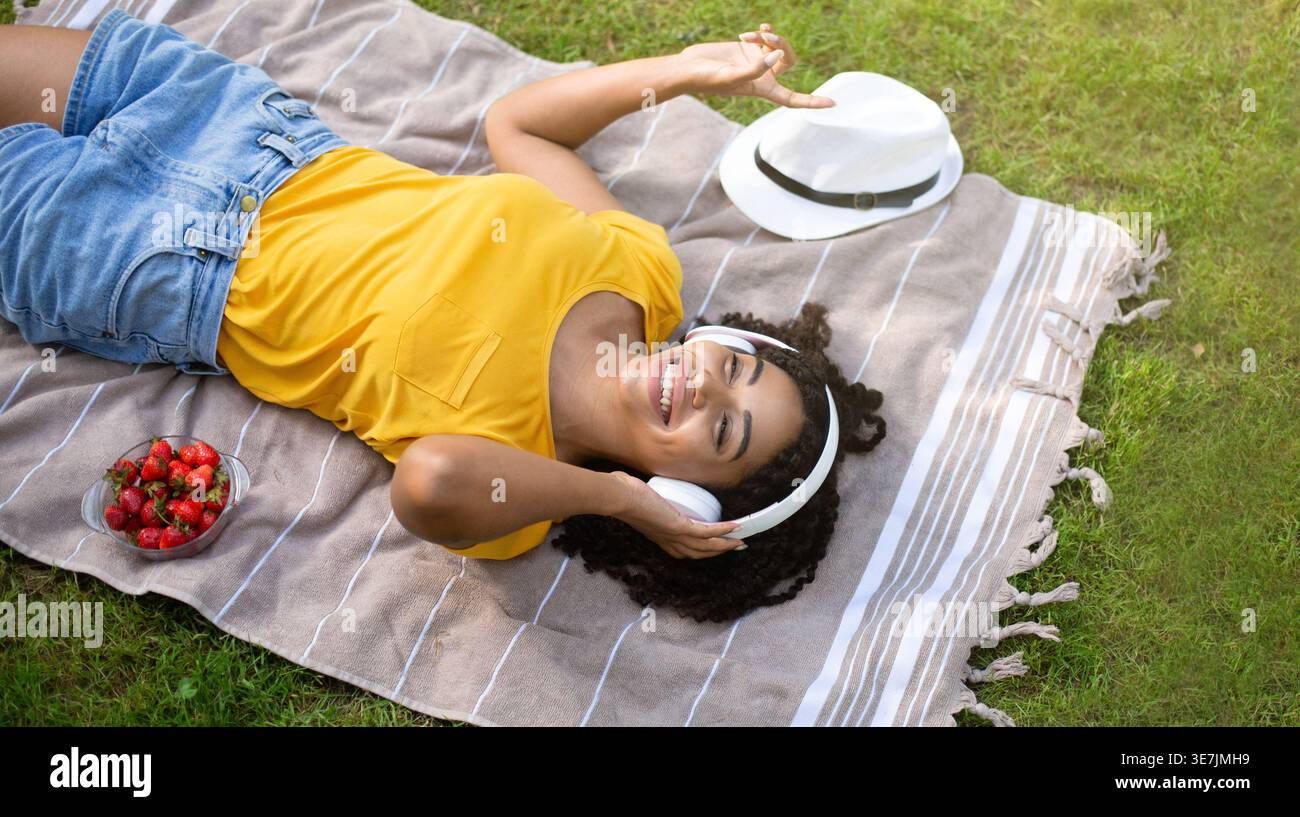 Sopra la vista di Black Woman con le cuffie si gode l'ascolto della musica mentre si è sdraiati sulla coperta del parco Foto Stock