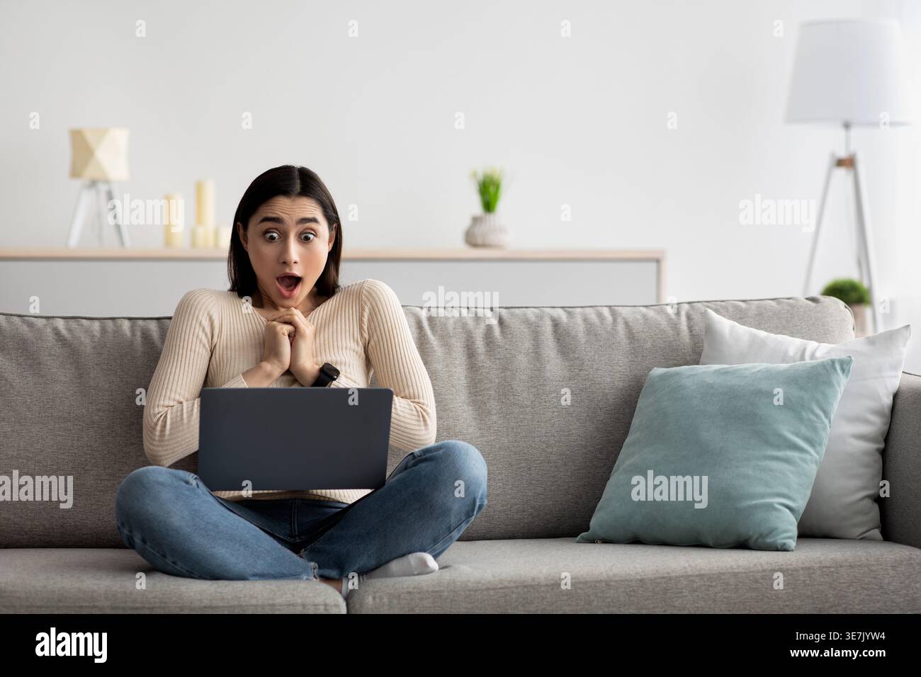 Una donna araba scioccata con le mani strette che guarda lo schermo di un laptop seduto con le gambe incrociate sul divano nel soggiorno di casa Foto Stock