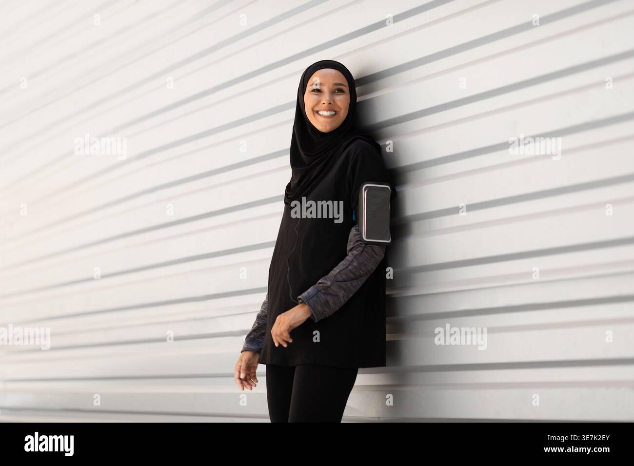 Atleta sorridente del Medio Oriente che indossa un hijab nero appoggiato su un roller shutter mentre è in piedi per strada durante il giorno Foto Stock