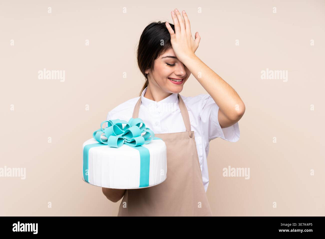 Pasticciera ispanica che tocca la fronte con gli occhi chiusi tenendo la torta in piedi sullo sfondo beige in studio Foto Stock