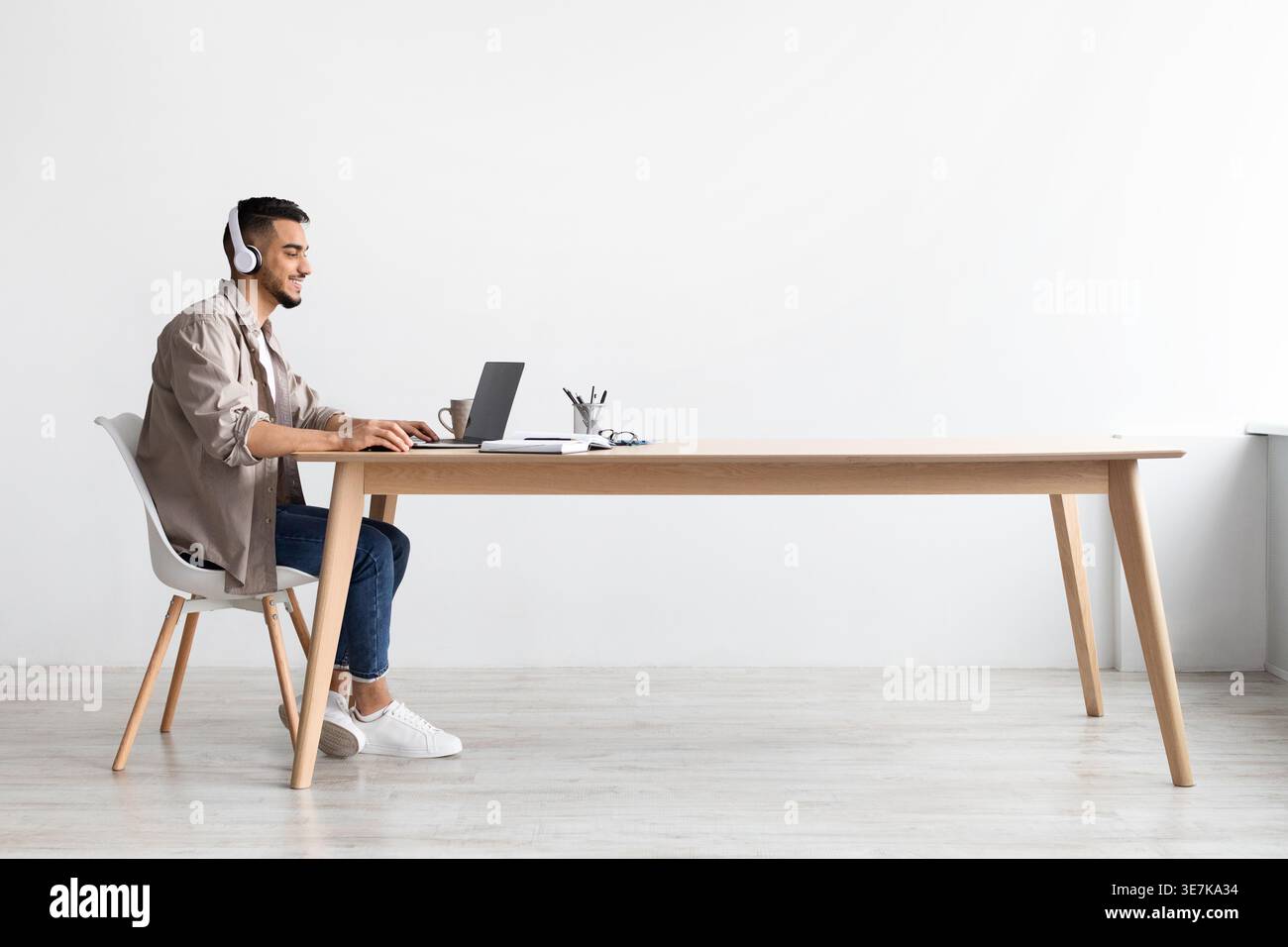Allegro uomo mediorientale che indossa le cuffie mentre lavora con un laptop seduto al tavolo in ufficio Foto Stock