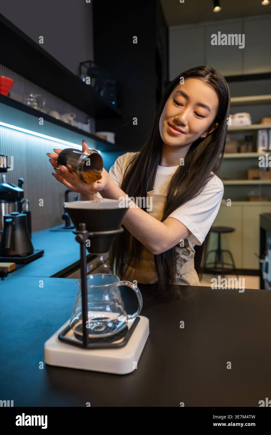 Sorridente lavoratrice asiatica del caffè che aggiunge caffè macinato all'imbuto della macchina del caffè con filtro nel caffè Foto Stock