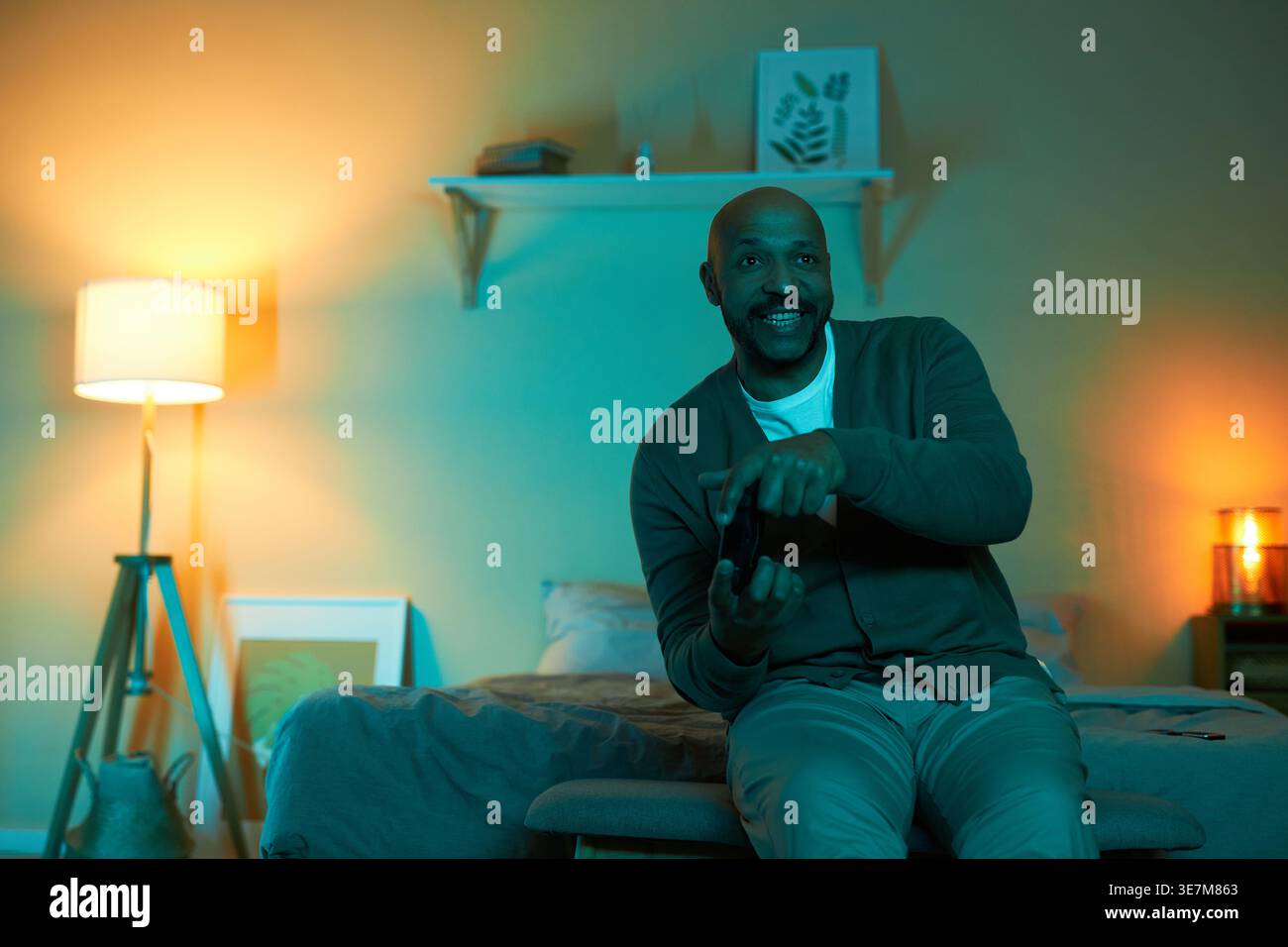 Uomo a corsa mista che tiene in mano il controller mentre gioca a vide in camera da letto di notte Foto Stock
