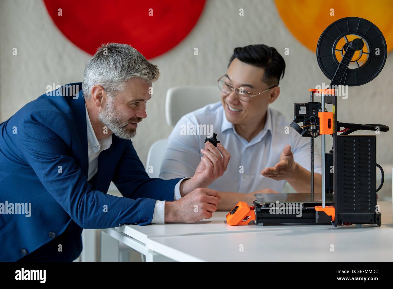 Ingegneri industriali maschi asiatici e caucasici che esaminano il prodotto creato da una stampante 3D mentre sono seduti al tavolo e lavorano insieme in ufficio Foto Stock