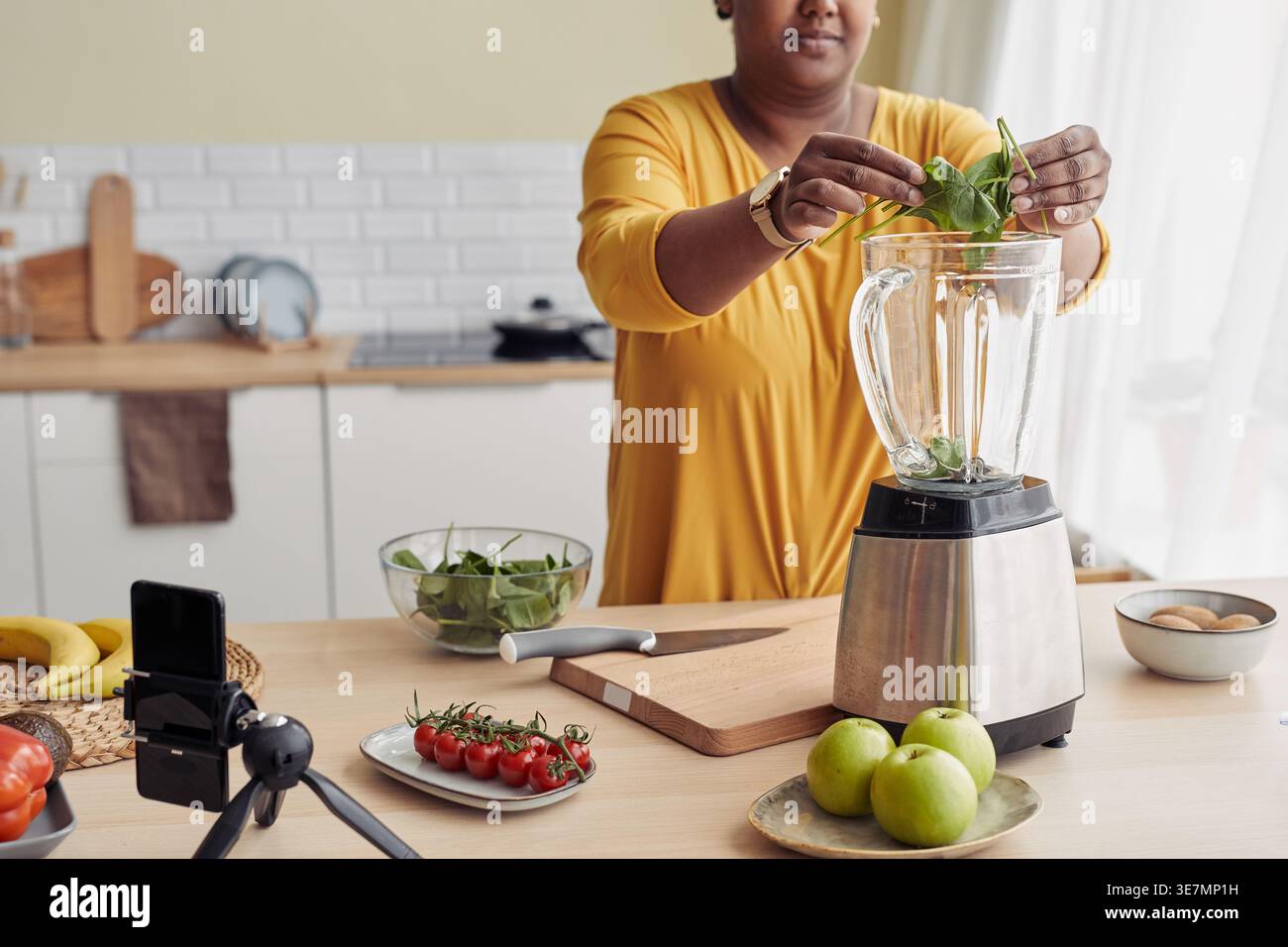 Donna nera sorridente e dalle dimensioni più grandi che mette gli spinaci nel frullatore per creare un frullato verde registrare video utilizzando lo smartphone su un treppiede in piedi al tavolo in kit... Foto Stock