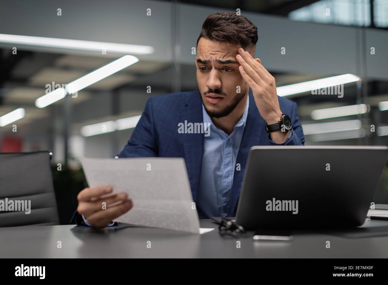 Un uomo d'affari arabo stressato leggendo un documento e toccando il tempio seduto alla scrivania con un portatile in ufficio Foto Stock
