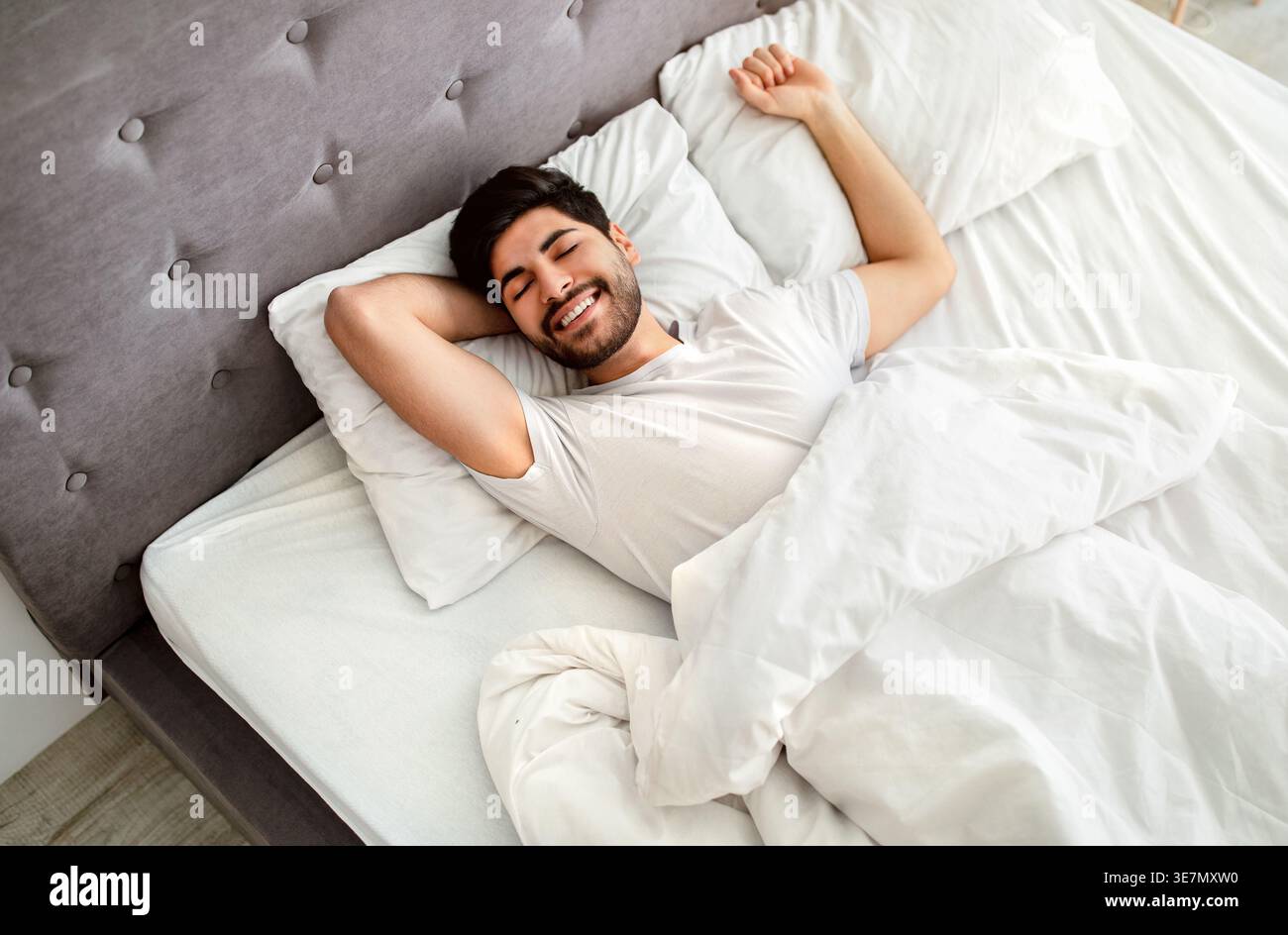 Uomo arabo sorridente con la barba che indossa una camicia bianca che allunga le braccia con gli occhi chiusi mentre è sdraiato sul letto in camera da letto di casa Foto Stock