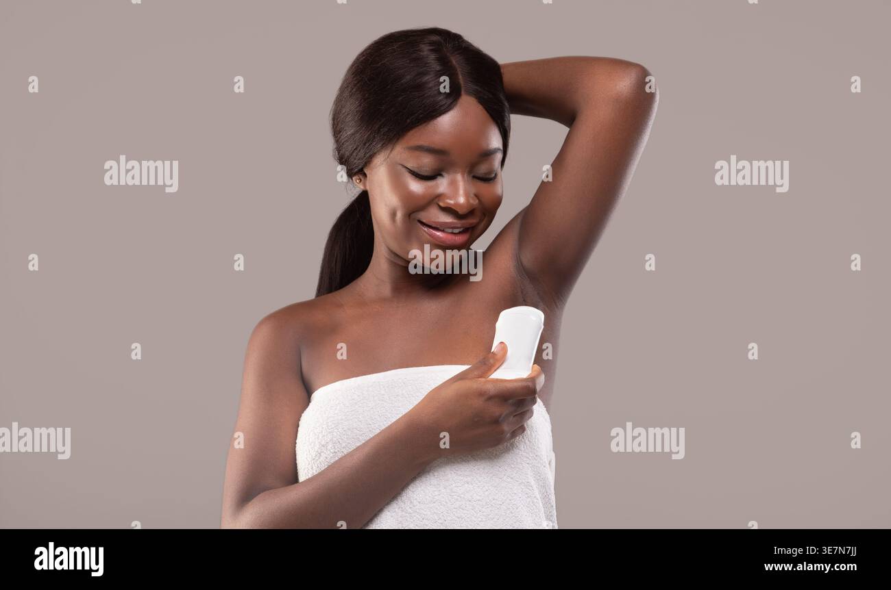 Donna nera avvolta in un asciugamano con deodorante mentre si trova su uno sfondo marrone rosato in studio Foto Stock