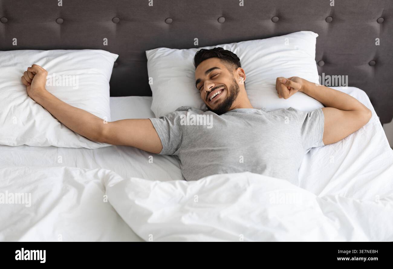 Uomo arabo che allunga le braccia mentre sdraiato sul letto nella camera da letto di casa Foto Stock