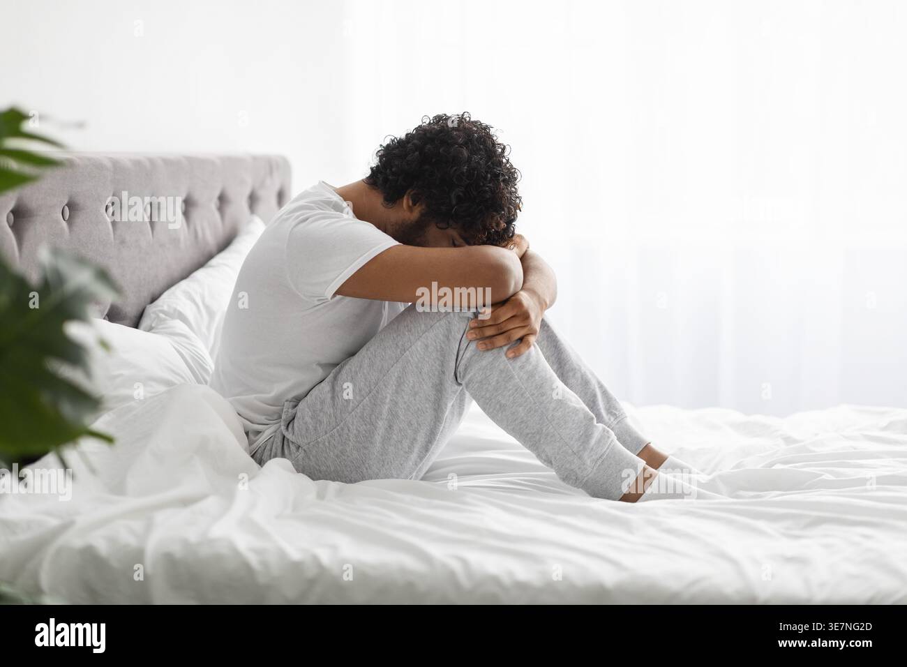 Uomo indiano depresso con capelli ricci appoggiati sul viso sulle ginocchia seduto sul letto in camera da letto Foto Stock