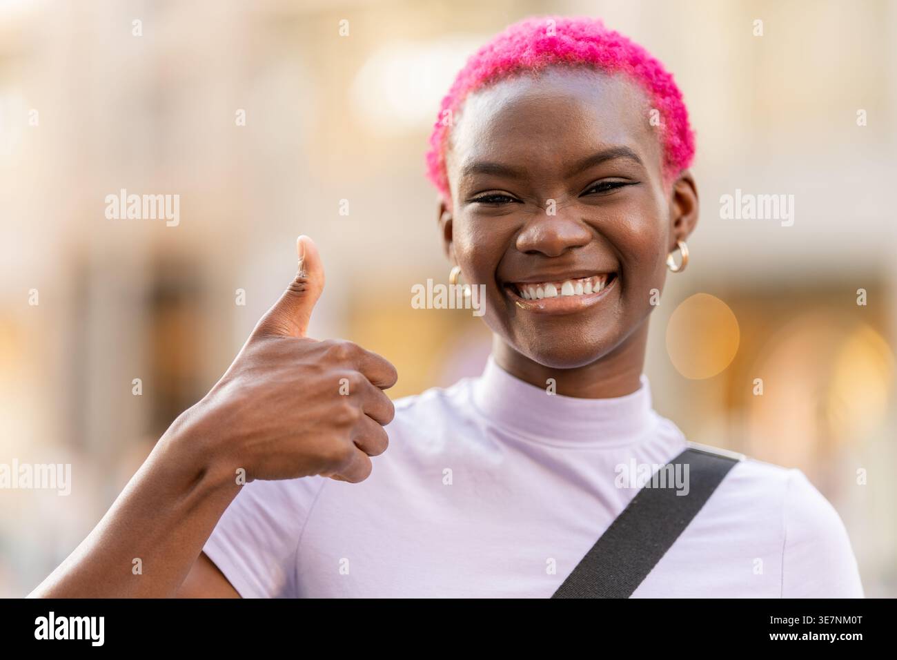 Donna nera sorridente con i capelli tinti corti che fanno i pollici in piedi sulla strada della città durante il giorno Foto Stock