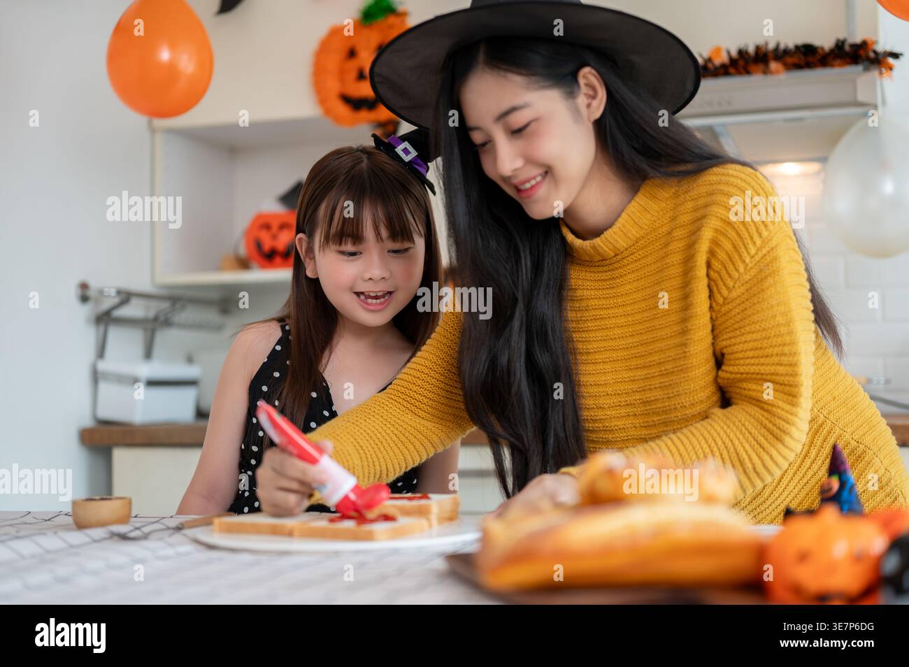 Madre e figlia asiatica sorridenti che indossano cappelli da strega che preparano il cibo per Halloween mentre sono seduti a tavola in cucina a casa Foto Stock