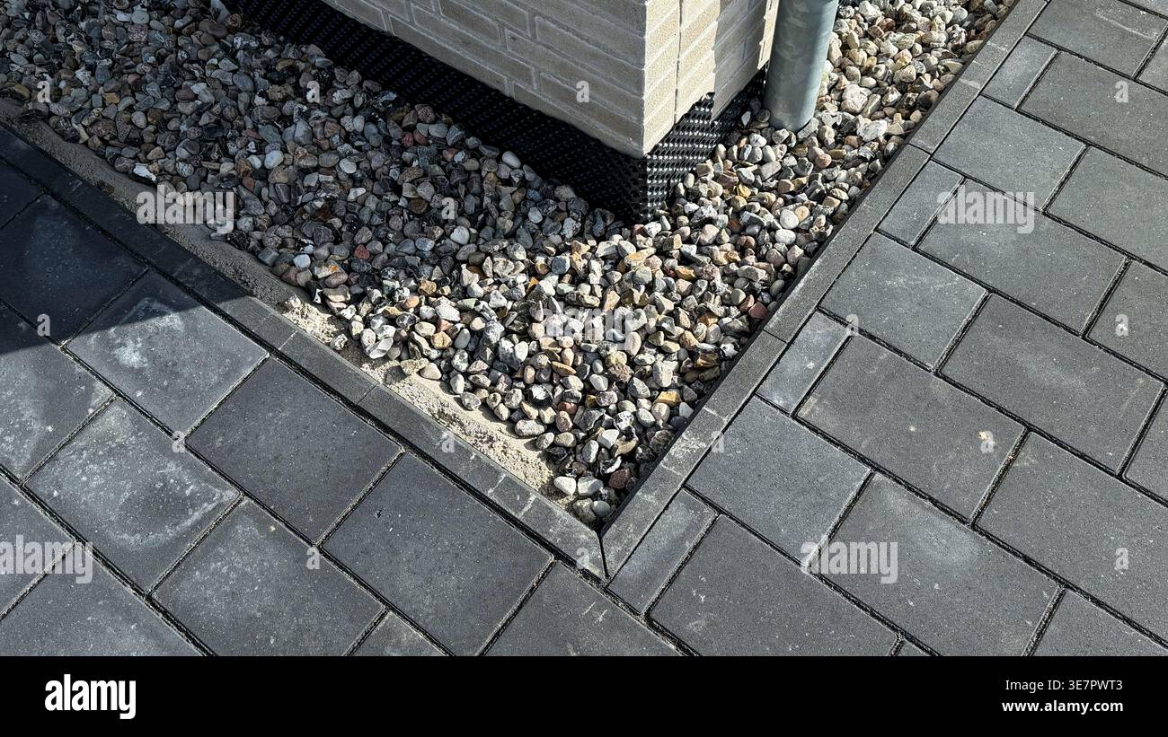 Vista ravvicinata di un angolo di un patio pavimentato con pietre grigie a incastro e ghiaia decorativa che circonda una colonna di mattoni con un tubo di drenaggio Foto Stock