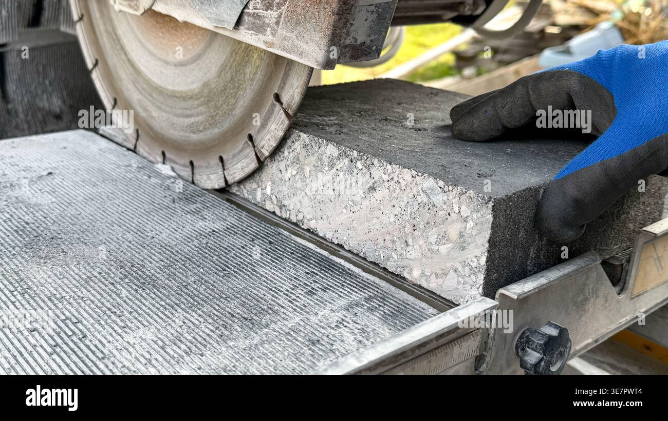 Lavoratore che utilizza una sega per calcestruzzo per tagliare un blocco di pietra su una piattaforma di taglio all'aperto, con uno sfondo sfocato di materiali e utensili da costruzione Foto Stock