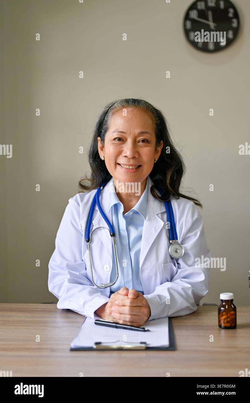 Medico asiatico anziano con le mani strette seduto a tavola in clinica Foto Stock