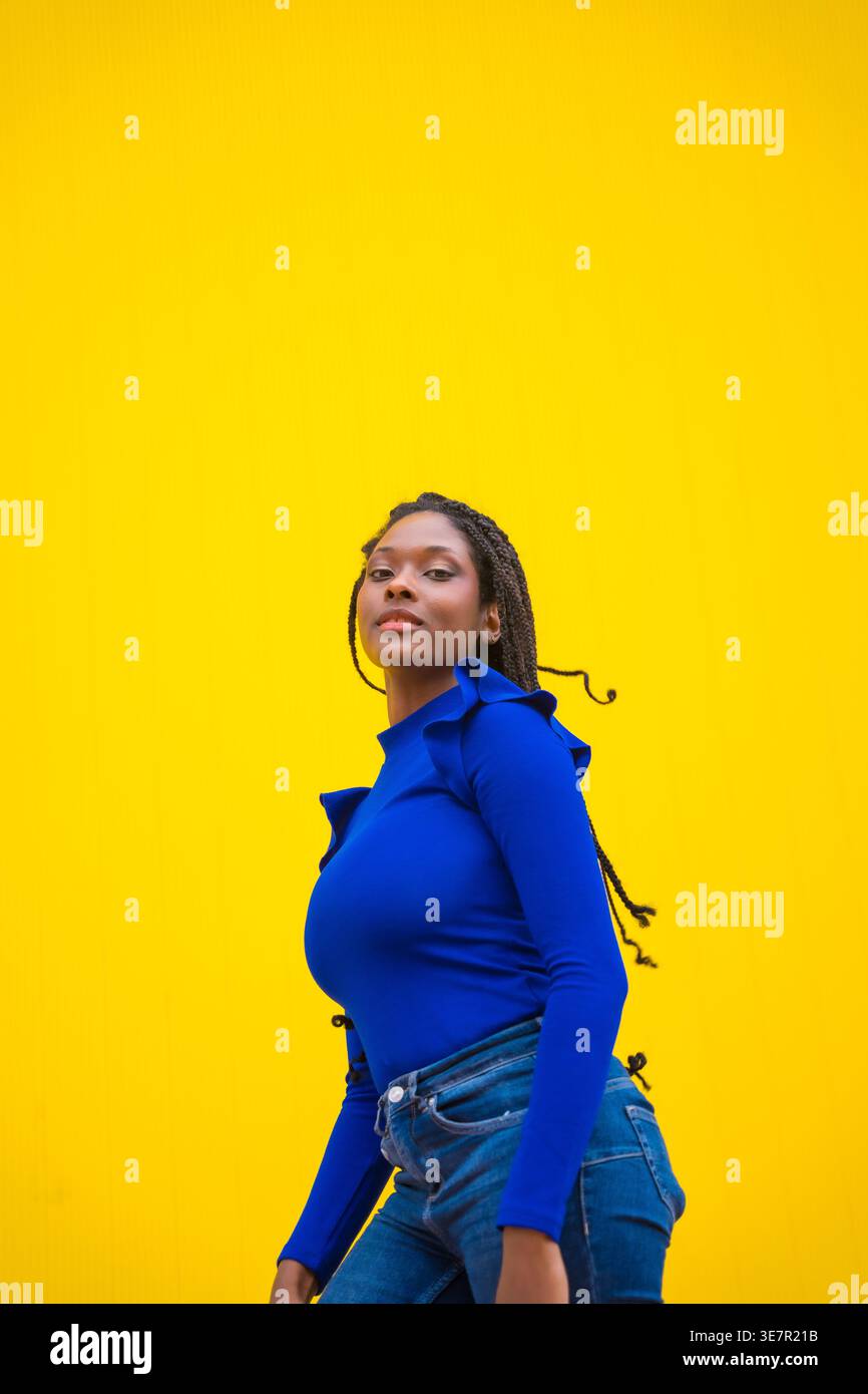 Donna nera con trecce lunghe che indossano maniche lunghe blu e jeans su sfondo giallo in studio Foto Stock
