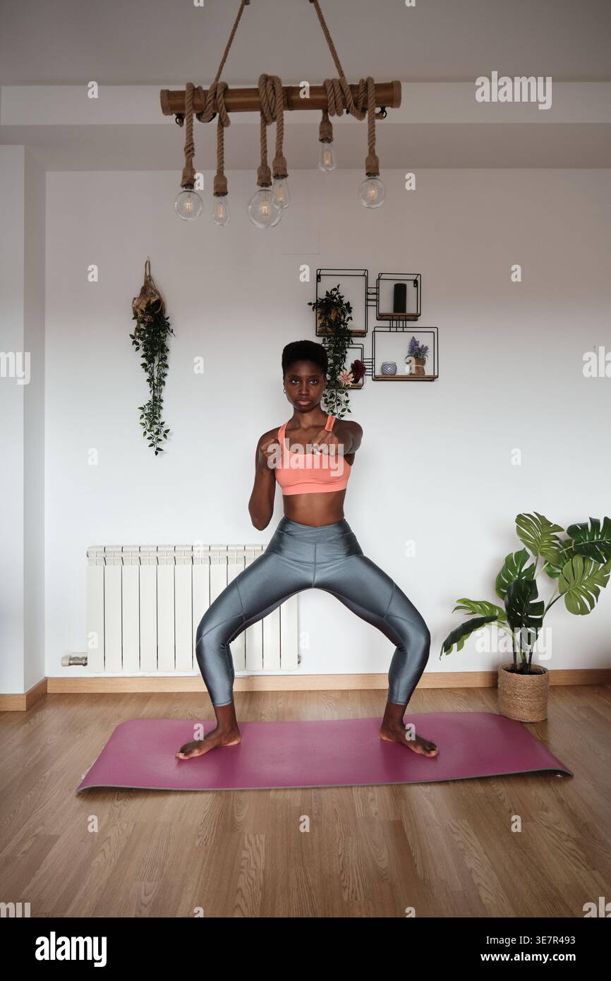 Donna nera con capelli corti in abbigliamento fitness che fa yoga in piedi con gambe piegate separate sul tappetino a casa Foto Stock