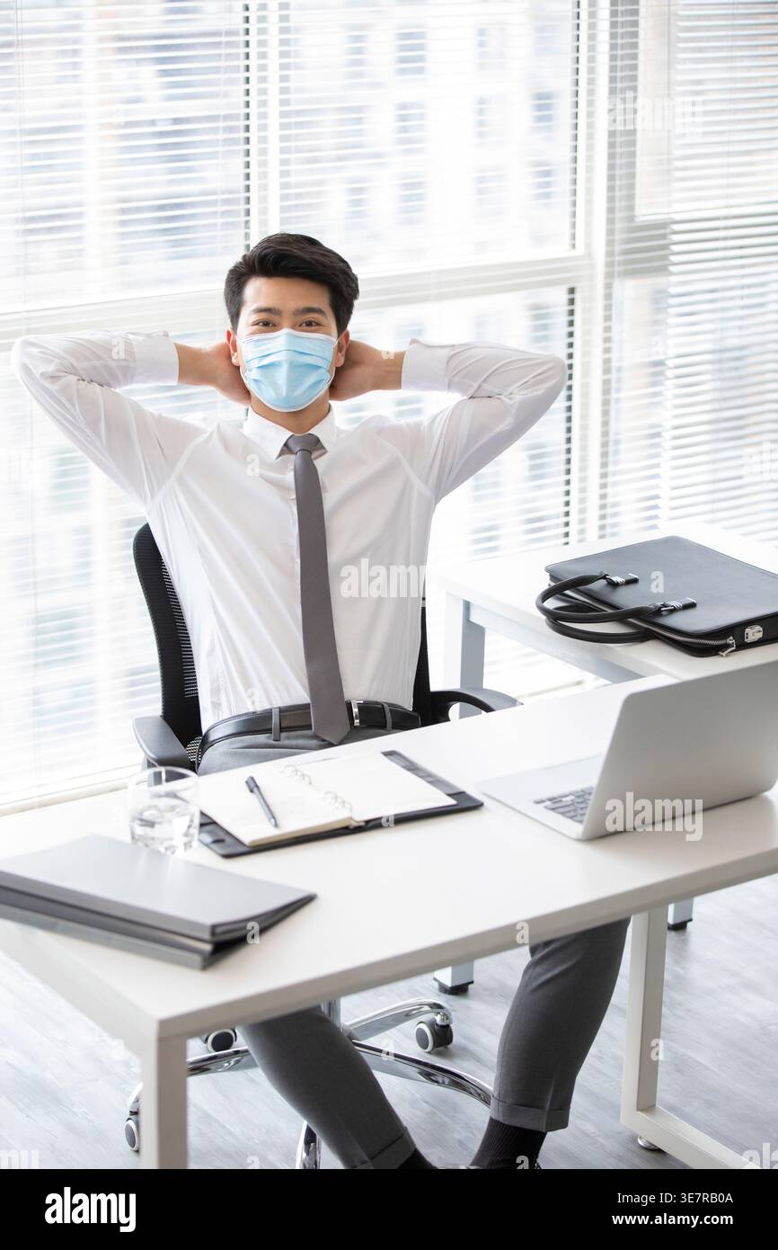 Sorridente uomo d'affari cinese che indossa la maschera seduta alla sua scrivania con le mani dietro la testa in ufficio Foto Stock